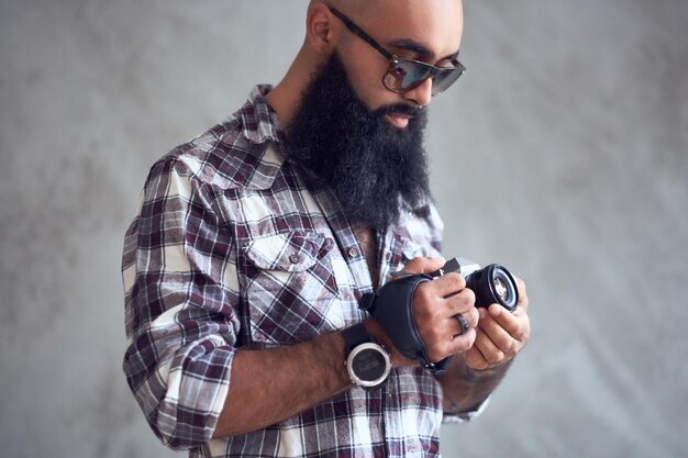 Un fotógrafo aficionado hipster barbudo con tatuajes en los brazos, vestido con una camisa de lana sostiene una cámara réflex digital compacta sobre fondo gris.