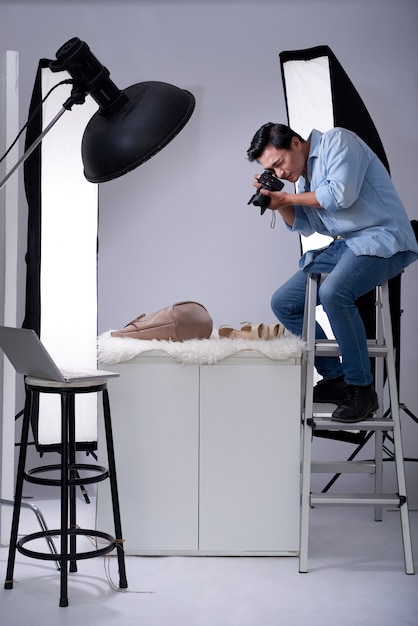 Fotógrafo asiático sentado en la escalera en el estudio con cámara y tomando fotos de artículos de moda