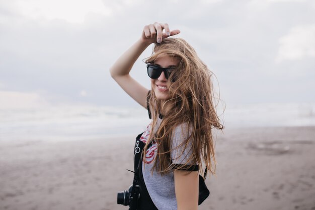 Fotógrafo de glamour sonriendo en día ventoso. Tiro al aire libre de elegante chica divertida que expresa felicidad mientras posa en la playa con la cámara.