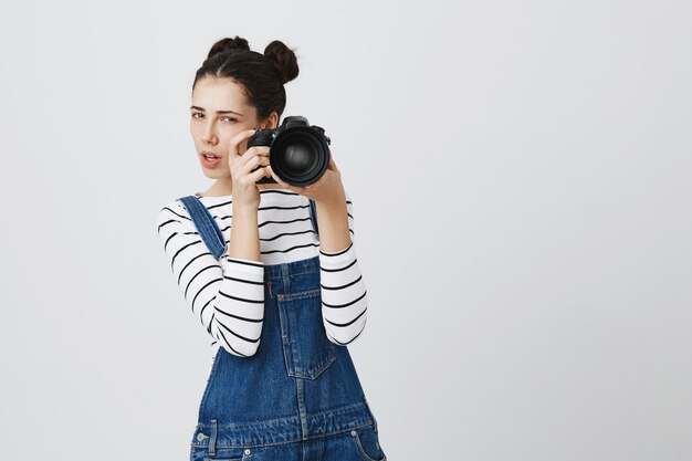 Fotógrafo de niña linda enfocada tomando fotografías en la cámara, mirando a su modelo pensativo