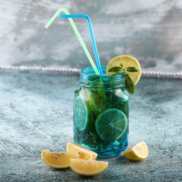 Un frasco de mojito azul con limón y menta sobre fondo brillante con tubos amarillos y azules.