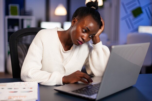 Freelancer africano agotado descansando la cabeza en la mano frente a la computadora portátil trabajando horas extras en la oficina de la empresa de puesta en marcha