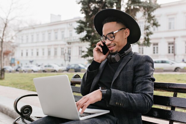 Freelancer africano complacido hablando por teléfono y escribiendo en el teclado. Foto al aire libre de estudiante internacional en traje negro usando laptop en la naturaleza.