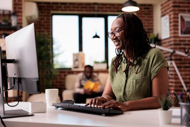 Freelancer afroamericano que trabaja escribiendo a distancia y mirando la pantalla de la computadora mientras su novio se relaja. Programador sonriente usando pc para chatear con amigos mientras el compañero de cuarto lee un libro.