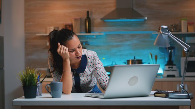 Freelancer agotado durmiendo frente a la computadora portátil trabajando desde la cocina de casa a altas horas de la noche. Empleado estresado que usa la red inalámbrica de tecnología moderna haciendo horas extras cerrando los ojos y durmiendo en la mesa.
