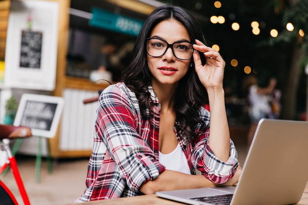 Freelancer bastante femenina lleva gafas de moda posando en la ciudad borrosa. Elegante chica de pelo negro usando laptop en buen día.