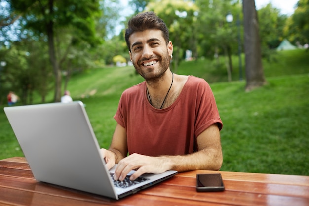 Freelancer guapo trabajando a distancia, sentarse en un banco del parque con una computadora portátil, conectar wifi