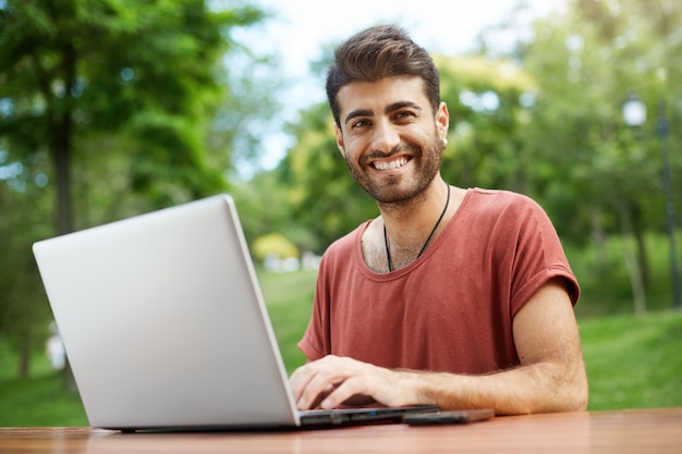 Freelancer guapo trabajando a distancia, sentarse en un banco del parque con una computadora portátil, conectar wifi