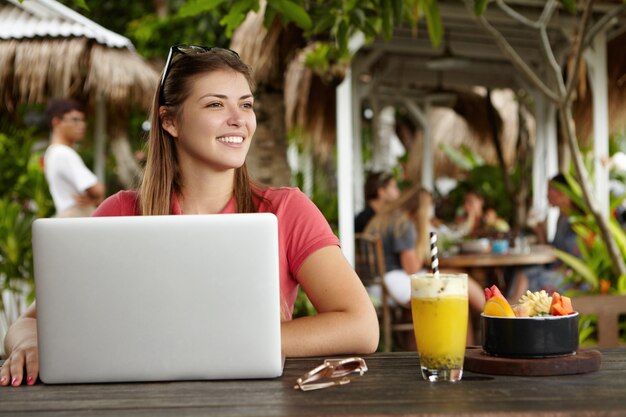 Freelancer hermosa joven vestida casualmente usando una computadora portátil genérica para trabajo remoto, descansando en un restaurante al aire libre