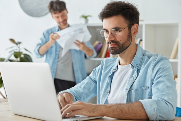 Freelancer hombre barbudo trabaja en computadora portátil, información de teclados, piensa en ganancias