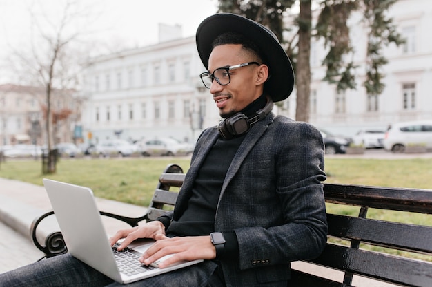 Freelancer joven hermoso que trabaja con la computadora en el parque. Retrato al aire libre de alegre chico africano con sombrero estudiando con un portátil en un banco.