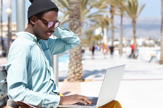 Freelancer joven de moda con ropa elegante y accesorios sentado en un banco