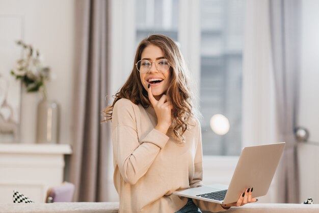 Freelancer mujer atractiva de buen humor disfrutando de sesión de fotos en interiores con ordenador portátil