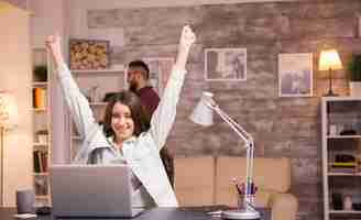 Foto gratuita freelancer mujer emocionada con las manos levantadas después de leer una buena noticia en la computadora portátil. novio hablando por teléfono en segundo plano.