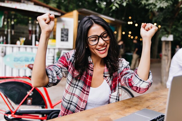 Freelancer mujer emocionada que trabaja en la cafetería al aire libre. Retrato de niña morena emocional sentada en la calle con portátil.