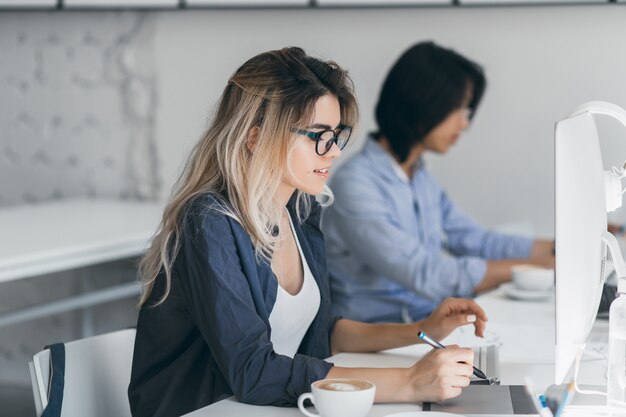 Freelancer mujer ocupada con cabello largo trabajando con tableta y tomando café. Retrato de interior de estudiante japonés concentrado usando computadora en el aula.