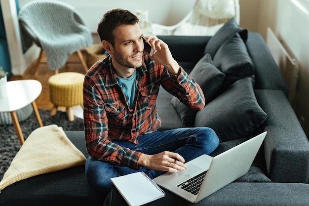 Freelancer sonriente hablando por teléfono móvil mientras trabaja en una computadora en la sala de estar.