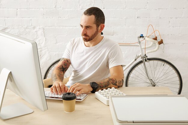 Freelancer tatuado barbudo en camiseta blanca en blanco trabaja en su computadora en casa frente a una pared de ladrillo y una bicicleta vintage estacionada, horario de verano