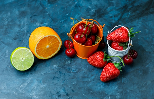 Fresas y cerezas en mini cubos con naranja, lima plana sobre un fondo azul sucio