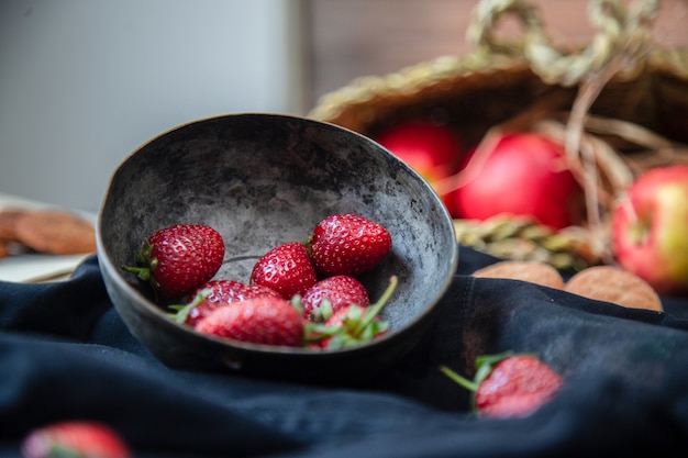 Fresas dentro del tazón de fuente, galletas y cesta de la manzana en una estera negra, cesta borrosa.