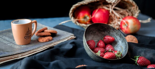 Fresas dentro del tazón, galletas, taza y cesta de manzana sobre una estera negra.