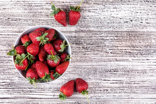 Fresas planas en un tazón y otras alrededor en una mesa de madera clara