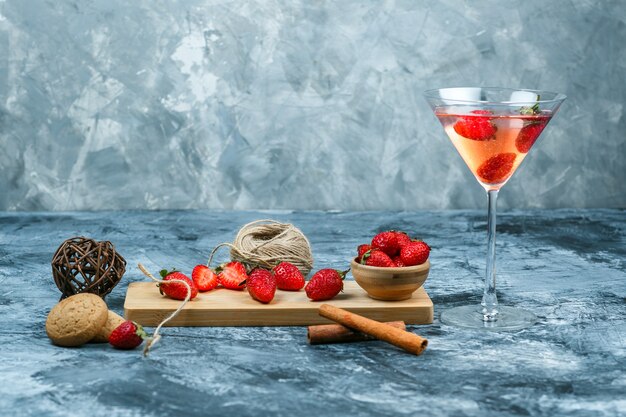 Fresas de primer plano y un cuchillo en la tabla de cortar con una copa de cóctel, ovillo, un tazón de fresas y galletas sobre fondo de mármol azul oscuro y gris. espacio libre horizontal para su texto