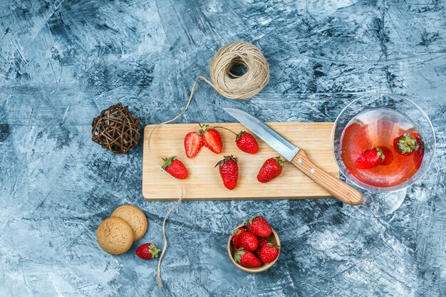 Fresas de primer plano y un cuchillo en la tabla de cortar con una copa de cóctel, ovillo, un tazón de fresas y galletas sobre fondo de mármol azul oscuro y gris. espacio libre horizontal para su texto