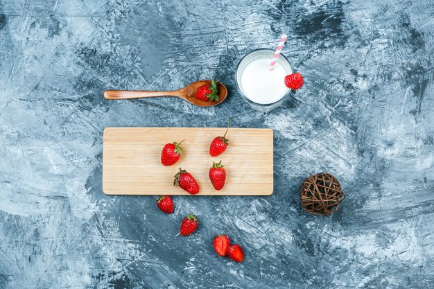 Fresas de vista superior en tabla de cortar con leche, ovillo y una cuchara de madera sobre superficie de mármol azul oscuro. horizontal