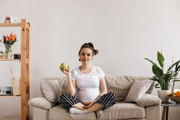 Foto gratuita fresca joven embarazada en camiseta blanca y pantalones a rayas tiene manzana verde