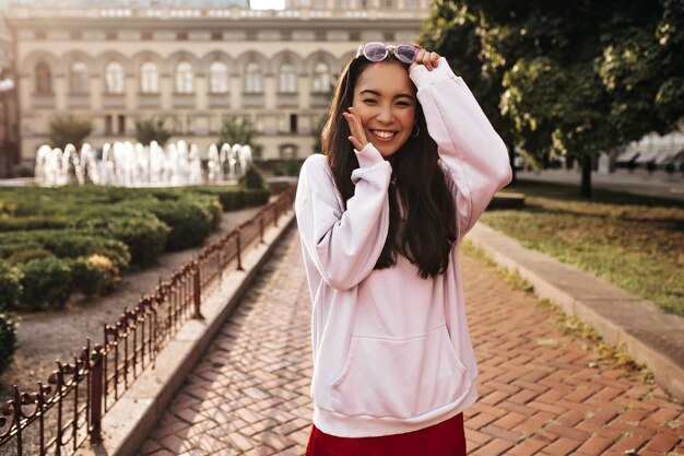 Fresca joven morena con sudadera con capucha rosa, falda de seda roja sonríe sinceramente, se ve feliz y se quita las gafas de sol afuera