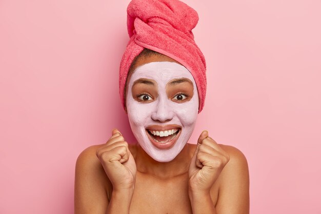 Frescura, ducha, concepto de autocuidado. Señora étnica llena de alegría levanta los puños cerrados, sonríe positivamente, hace tratamientos de belleza después del baño, la cara cubierta con mascarilla hidratante de arcilla, cuerpo desnudo y saludable