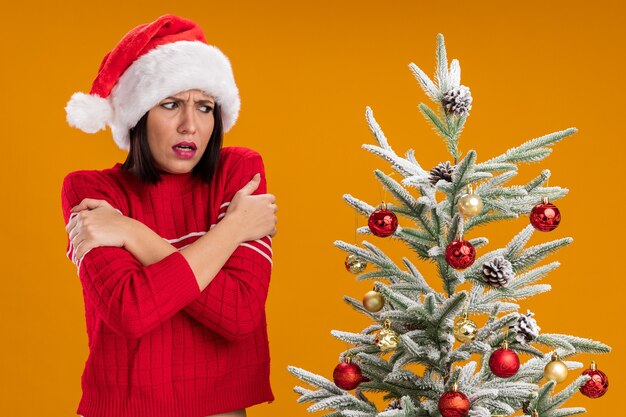 Fría niña con gorro de Papá Noel de pie cerca del árbol de navidad decorado mirándolo manteniendo las manos cruzadas sobre los brazos aislados en la pared naranja