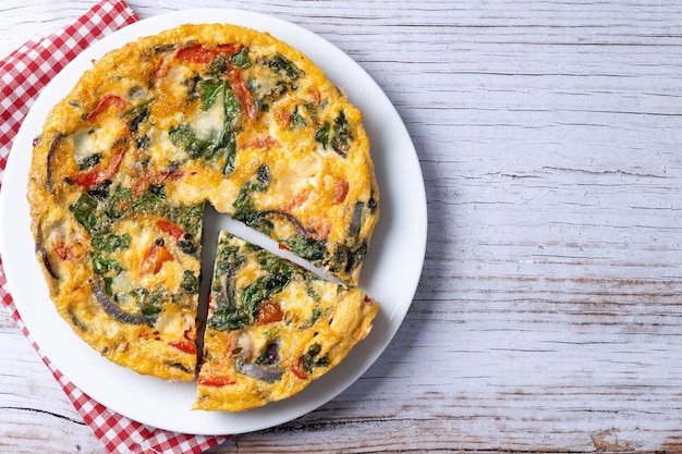 Frittata italiana hecha con espinacas, tomates, cebolla y pimientos sobre una mesa de madera blanca