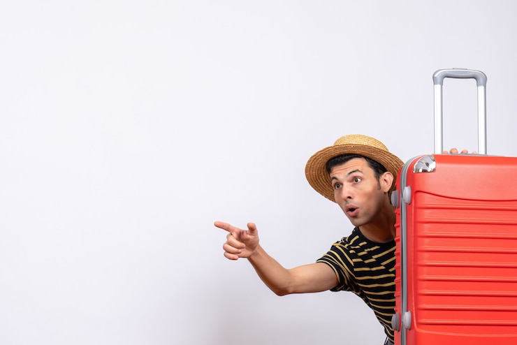 Photo Gratis front view young man with straw hat hiding behind red suitcase pointing at something