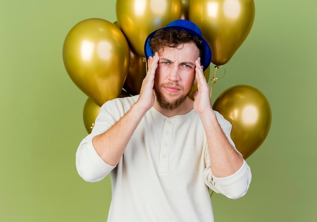 Frunciendo el ceño joven apuesto chico de fiesta eslavo con sombrero de fiesta de pie delante de globos mirando al frente manteniendo las manos en la cabeza que sufren de dolor de cabeza aislado en la pared verde oliva