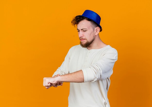 Frunciendo el ceño joven apuesto chico de fiesta eslavo con sombrero de fiesta que señala el dedo en la muñeca finge mirar el reloj aislado sobre fondo naranja con espacio de copia