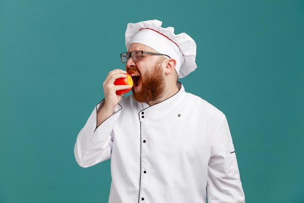 Frunciendo el ceño joven chef masculino con gafas uniformes y gorra mordiendo manzana con los ojos cerrados aislado sobre fondo azul.