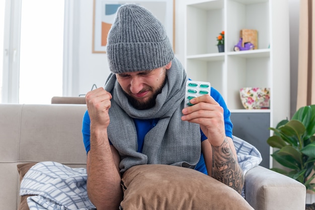 Foto gratuita frunciendo el ceño joven enfermo con bufanda y gorro de invierno sentado en el sofá en la sala de estar con una almohada en las piernas sosteniendo un paquete de cápsulas haciendo un gesto de sí con los ojos cerrados