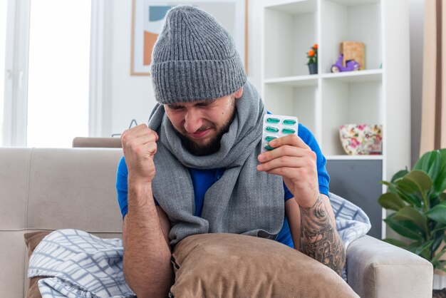 Frunciendo el ceño joven enfermo con bufanda y gorro de invierno sentado en el sofá en la sala de estar con una almohada en las piernas sosteniendo un paquete de cápsulas haciendo un gesto de sí con los ojos cerrados