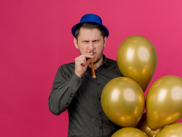 Frunciendo el ceño a un joven fiestero con sombrero azul sosteniendo globos que soplan silbato aislado en rosa