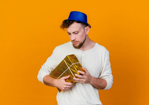 Frunciendo el ceño joven guapo chico partido eslavo con sombrero de fiesta mirando hacia abajo sosteniendo una caja de regalo aislada sobre fondo naranja con espacio de copia
