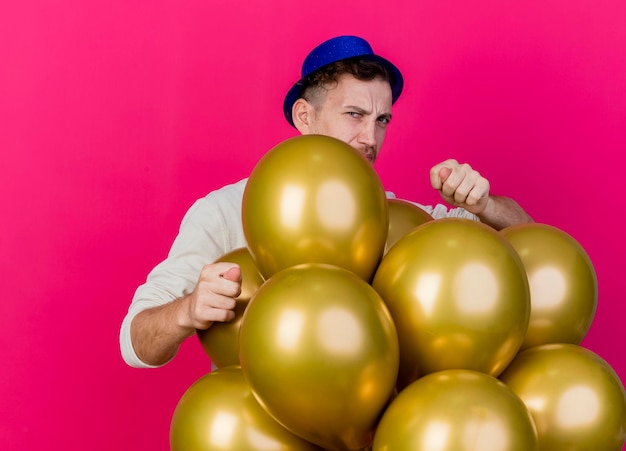 Frunciendo el ceño joven guapo chico partido eslavo con sombrero de fiesta de pie detrás de globos mirando al frente haciendo signo de higo aislado en la pared rosa