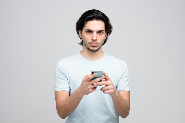 frunciendo el ceño joven guapo sosteniendo el teléfono móvil con ambas manos mirando a la cámara aislada sobre fondo blanco