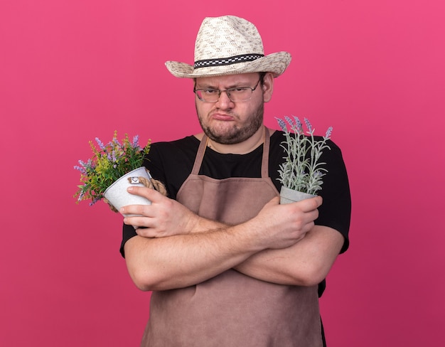 Frunciendo el ceño joven jardinero con sombrero de jardinería sosteniendo y cruzando flores en macetas aisladas en la pared rosa