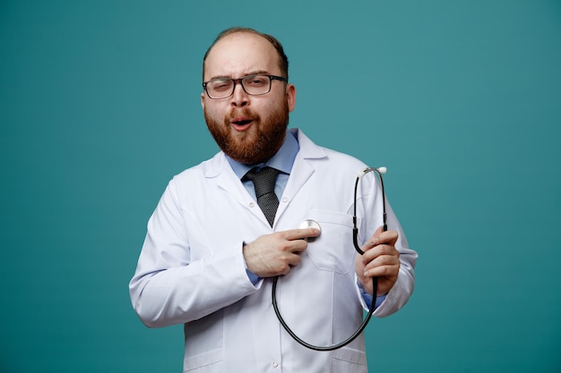 Frunciendo el ceño joven médico masculino con gafas abrigo médico mirando a la cámara poniendo estetoscopio en el corazón aislado sobre fondo azul.