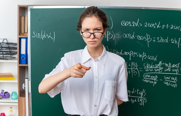 Frunciendo el ceño joven profesora de matemáticas con gafas de pie delante de la pizarra manteniendo la mano detrás de la espalda mirando y apuntando al frente en el aula
