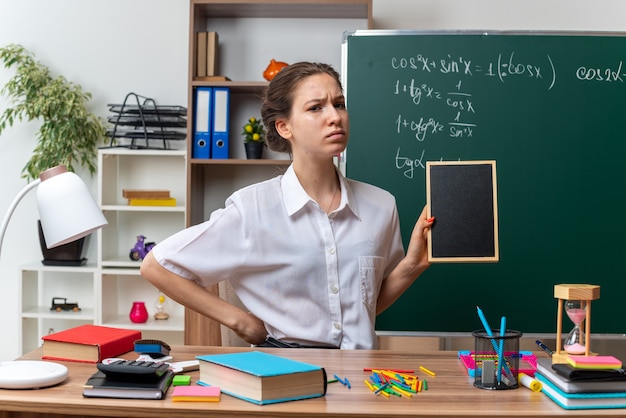 Frunciendo el ceño joven profesora de matemáticas sentada en un escritorio con útiles escolares sosteniendo una mini pizarra manteniendo la mano en la cintura mirando al frente en el aula