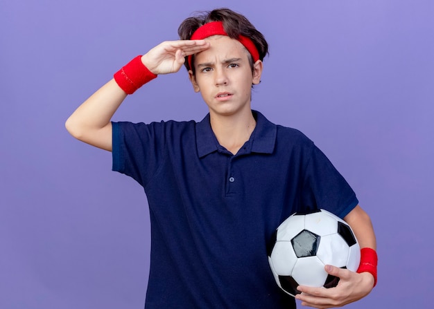 Fruncir el ceño joven apuesto deportista con diadema y muñequeras con aparatos dentales sosteniendo un balón de fútbol mirando al frente manteniendo la mano en la frente aislada en la pared púrpura