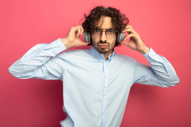 Fruncir el ceño joven apuesto hombre con gafas y auriculares mirando al frente agarrando auriculares aislados en la pared rosa